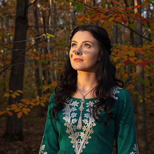 Photograph of a fair-skinned woman with long black hair, wearing a green embroidered top, standing in a sunlit autumn forest.