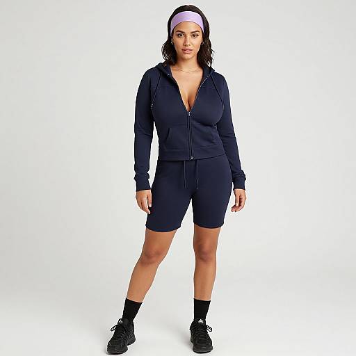 Photograph of a curvy woman with medium brown skin, black hair, wearing a navy zip-up hoodie, shorts, white headband, black socks