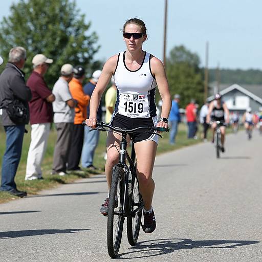 Photograph of a fit woman with dark hair in sunglasses, wearing a white and black racing tank top and shorts, cycling on a sunny road in a