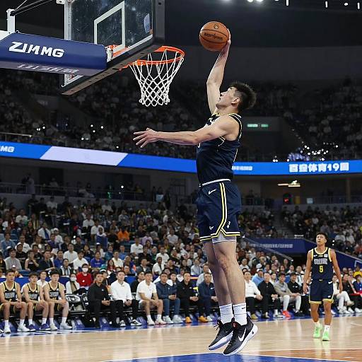 Photograph of a male basketball player in a navy uniform, mid-jump shot, under the hoop, in a packed arena with bright lights.