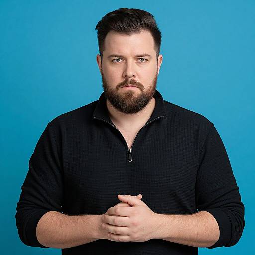 Photograph of a bearded, fair-skinned man with short dark hair, wearing a black zip-up sweater, against a bright blue background. He