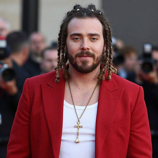 Photograph of a bearded man with dreadlocks wearing a red blazer, white shirt, and gold necklace, standing in front of a blurred crowd