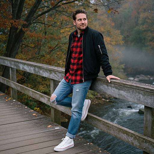 Photograph of a bearded man in a red plaid shirt, black jacket, ripped jeans, and white sneakers, leaning on a wooden bridge with