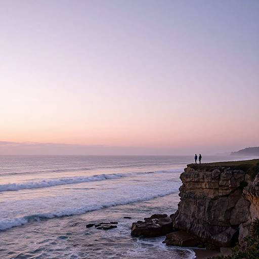 Photograph of two silhouetted figures standing on a rocky cliff at sunset, overlooking a serene ocean with gentle waves.