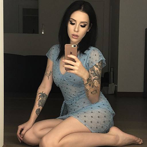Photograph of a tattooed woman with pale skin, black hair, and full lips, wearing a sheer blue polka dot dress, kneeling on the