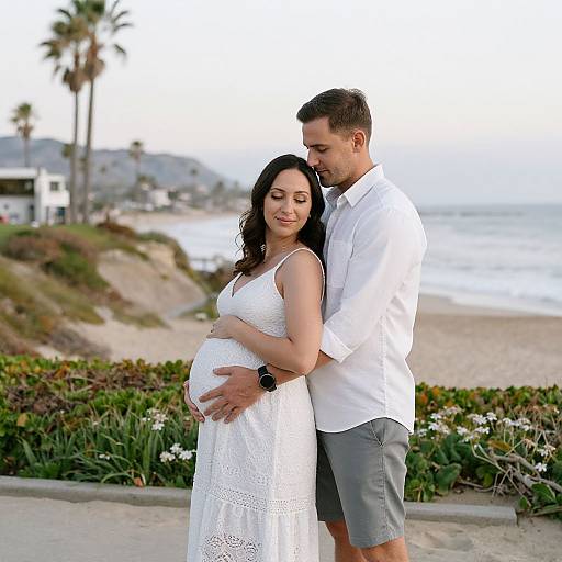Photograph of a pregnant couple standing on a beach; man in white shirt and gray shorts, woman in white lace dress, arms around each other,
