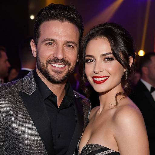 Photograph of a smiling couple at a red carpet event; the man with dark hair and beard wears a black and silver suit, the woman with dark