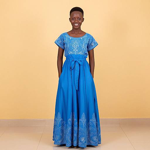 Photograph of a smiling Black woman with short hair, wearing a vibrant blue, patterned, short-sleeve, floor-length dress, standing against