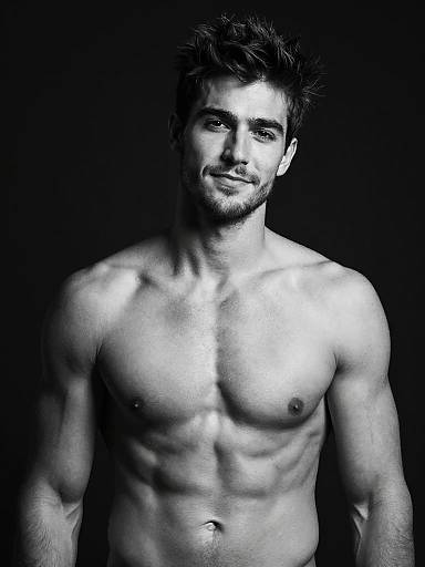Black-and-white photograph of a muscular, shirtless man with short dark hair and beard, standing against a dark background, highlighting his defined abs and chest