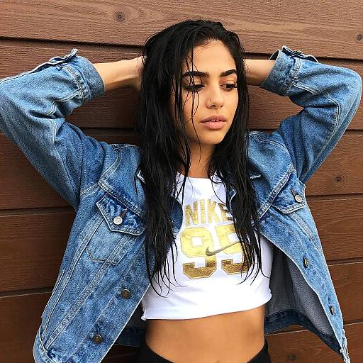 Photograph of a wet-haired, light-skinned woman with dark eyebrows, wearing a blue denim jacket over a white crop top with gold print, standing