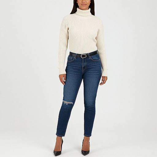Photograph of a woman in a white ribbed turtleneck, blue distressed jeans with a rip, and black high heels, standing against a white
