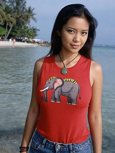 Photograph of an Asian woman with long black hair, wearing a red tank top with an elephant graphic and jeans, standing on a tropical beach with palm