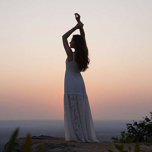 Silhouetted woman with long curly hair, wearing a white lace dress, raises arms against a pink and purple sunset sky.