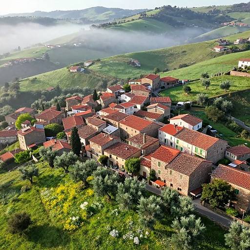 Aerial photograph of a picturesque, hillside Italian village with red-tiled roofs, surrounded by lush green fields and misty hills.