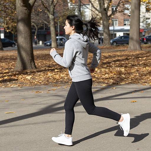 Photograph of a woman with black hair in a ponytail, jogging in a park wearing a gray hoodie, black leggings, and white sneakers, surrounded