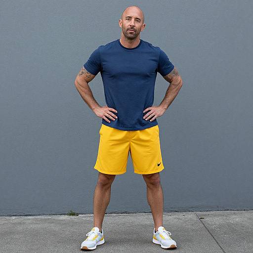 Photograph of a muscular, bald, tan-skinned man with a beard, wearing a navy t-shirt, yellow shorts, and white sneakers, standing