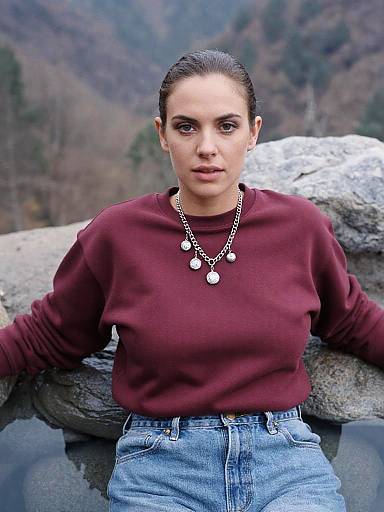 Photograph of a young woman with light skin and dark hair in a bun, wearing a maroon sweater, denim high-waisted jeans, and