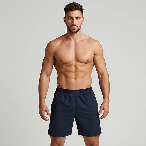 Photograph of a muscular, shirtless man with short dark hair and beard, wearing black shorts, standing against a plain white background.