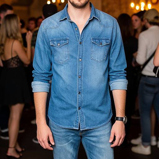 Photograph of a bearded man in a blue denim shirt and jeans, standing in a dimly lit, bustling social event.