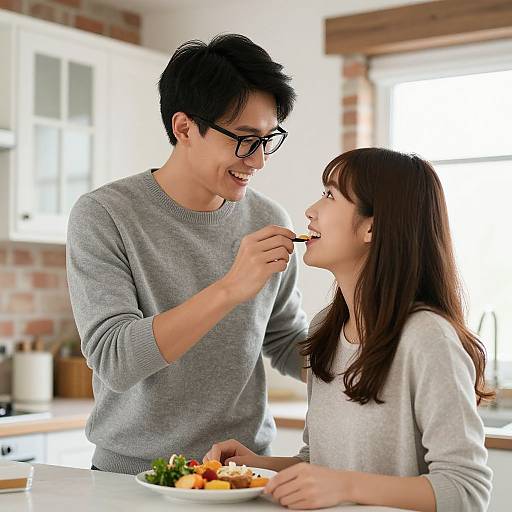 Photograph of an Asian couple sharing food in a bright kitchen; man with glasses, gray sweater, feeding woman in white sweater.