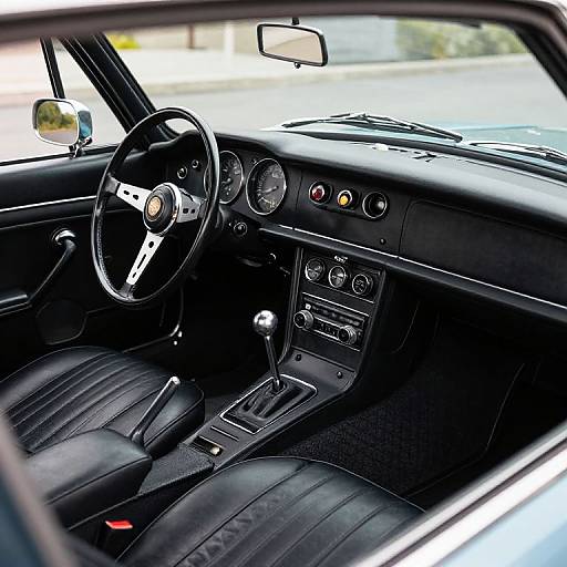 Photograph of a vintage car interior, showing a black steering wheel, analog dashboard, black leather seats, gear shift, and exterior sunlight.