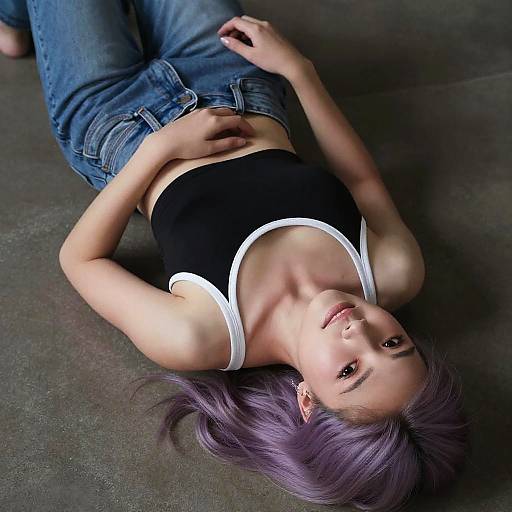 Photograph of a young woman with purple hair, lying on a dark floor, wearing a black tank top with white trim and blue jeans, looking up
