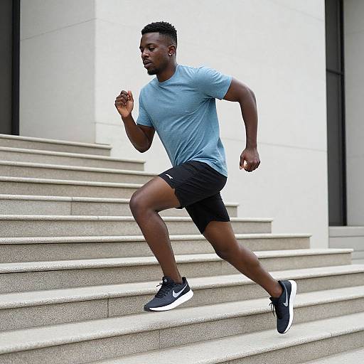 Photograph of a muscular Black man jogging up stone stairs, wearing a light blue t-shirt, black shorts, and black sneakers. Bright, minimalist background
