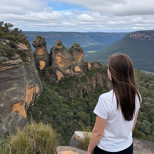 Photograph of a woman with long brown hair in a white t-shirt, standing on a rocky mountain overlook, gazing at rugged, forested cliffs