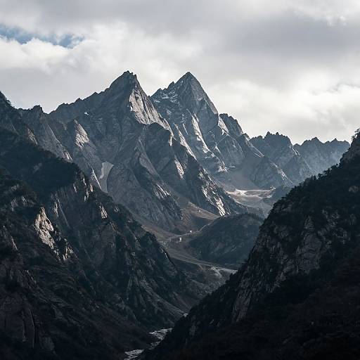 Photograph of a dramatic mountain range with jagged peaks, dark rocky slopes, and a bright, cloudy sky overhead. Shadows cast across the valley create