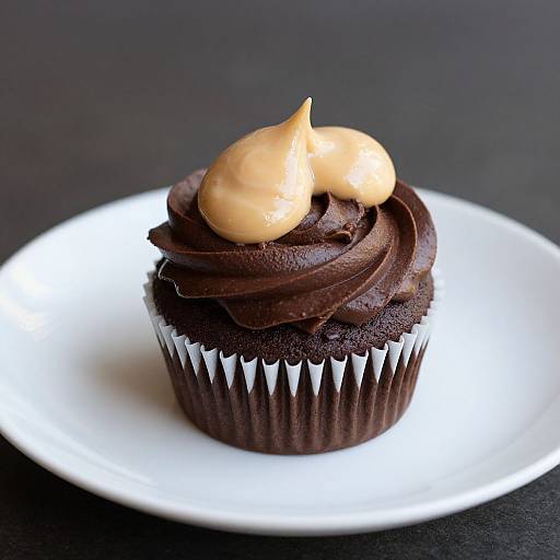 Photograph of a chocolate cupcake with dark, swirled frosting topped with two golden-brown caramel-like dollops, on a white plate against