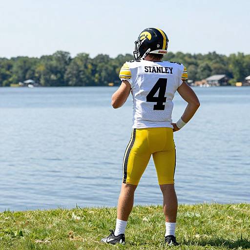 Photograph of a male football player in white and yellow uniform, numbered 4, standing on grassy bank, facing lake and trees.