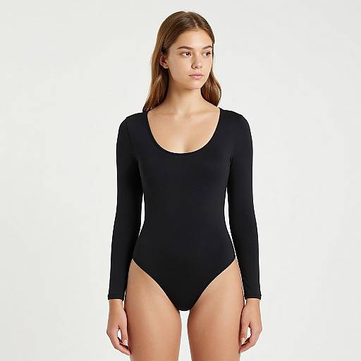 Photograph of a young woman with light skin and brown hair, wearing a black, long-sleeve, high-cut leotard, standing against