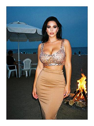 Photograph of a curvy woman with dark hair in a sparkly, low-cut, beige dress, standing on a beach at night by a fire