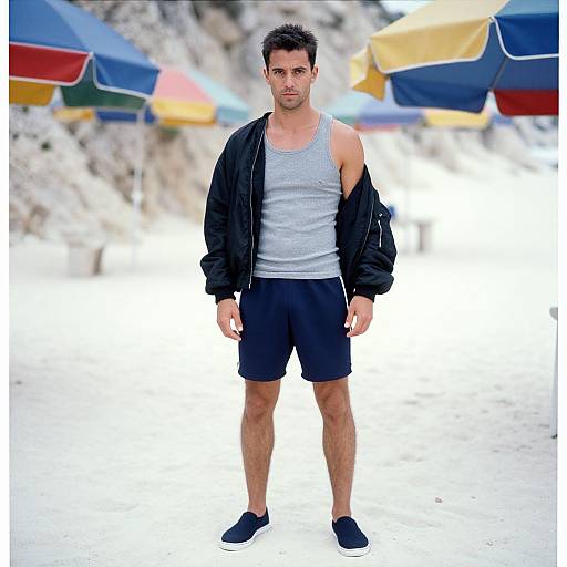 Photograph of a fit, dark-haired man in a gray tank top, black shorts, and black jacket, standing on a bright beach with colorful umb