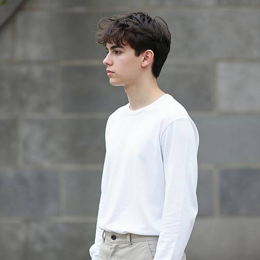 Photograph of a young man with fair skin, dark brown curly hair, wearing a white long-sleeve shirt and beige pants, standing in profile
