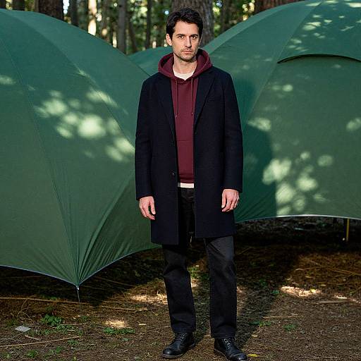 Photograph of a man with short dark hair, wearing a black coat, maroon hoodie, and black pants, standing in front of two green camping