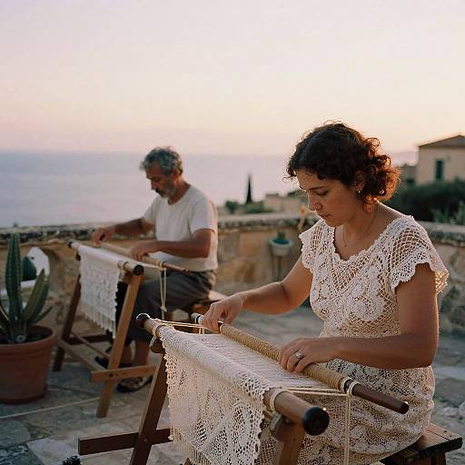 Photograph of two elderly artisans weaving lace on outdoor looms at sunset, with a coastal background and potted cactus.