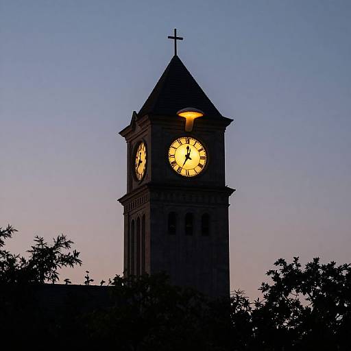 Photograph of a clock tower at dusk, illuminated clock face glowing yellow, silhouetted against a gradient blue to purple sky, with tree branches