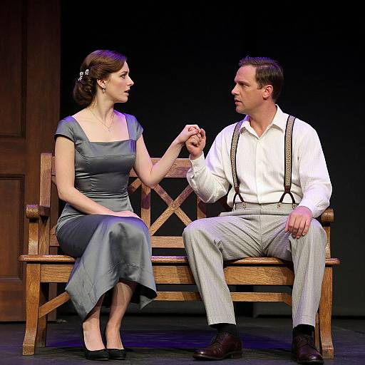 Photograph of a stage scene featuring a woman in a blue dress and a man in a white shirt and suspenders, seated on a wooden bench,