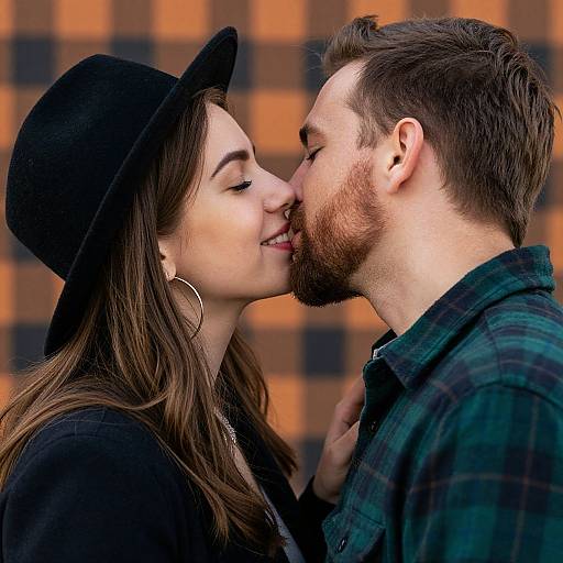 Photograph of a smiling couple sharing a kiss; woman in black hat and earrings, man in green plaid shirt, orange-checkered background.