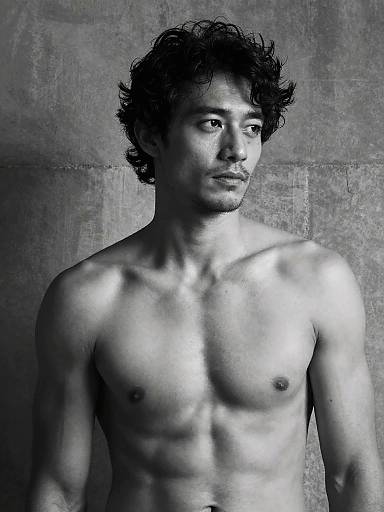 Black-and-white photograph of a shirtless, muscular Asian man with curly hair, serious expression, and smooth skin, against a textured gray background.