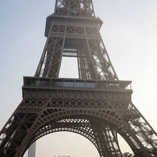 Photograph of the Eiffel Tower's upper structure, showcasing its intricate iron lattice, large archway, and bright sunlit background.