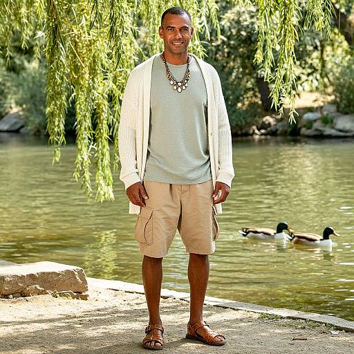 Photograph of a smiling, medium-built Black man with short hair, wearing a white shirt, beige shorts, necklace, and sandals, standing by a