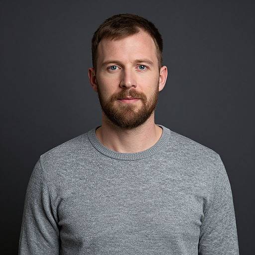 Photograph of a white man with short brown hair, blue eyes, and a full beard, wearing a gray, textured sweater, against a dark blue