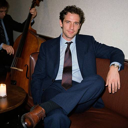 Photograph of a casually dressed man in a dark suit, light blue shirt, and dark tie, sitting on a brown couch, with a musician playing