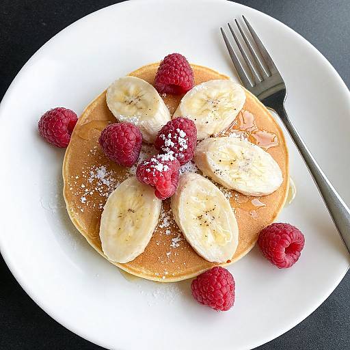 Photograph of a white plate with golden pancakes, sliced bananas, raspberries, and powdered sugar; a fork on the right.