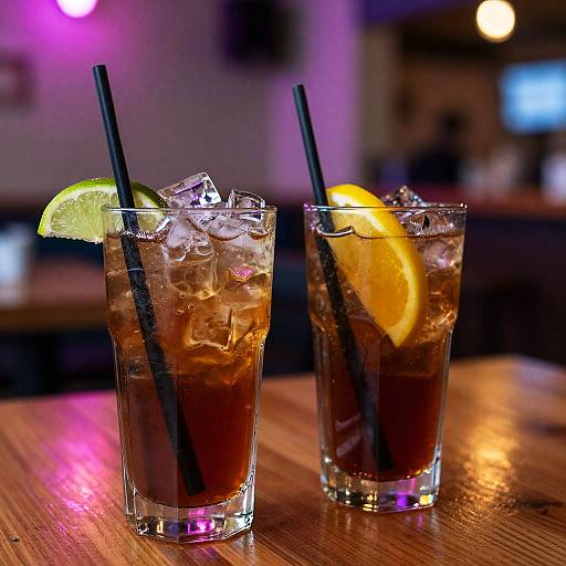 Photograph of two tall glasses with ice, dark mixed drinks, black straws, lime and orange slices, on a wooden bar table under purple and