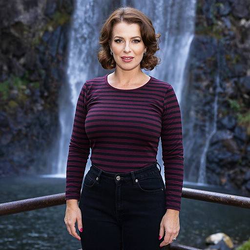 Photograph of a middle-aged woman with brown wavy hair, wearing a striped purple shirt and black pants, standing in front of a waterfall.