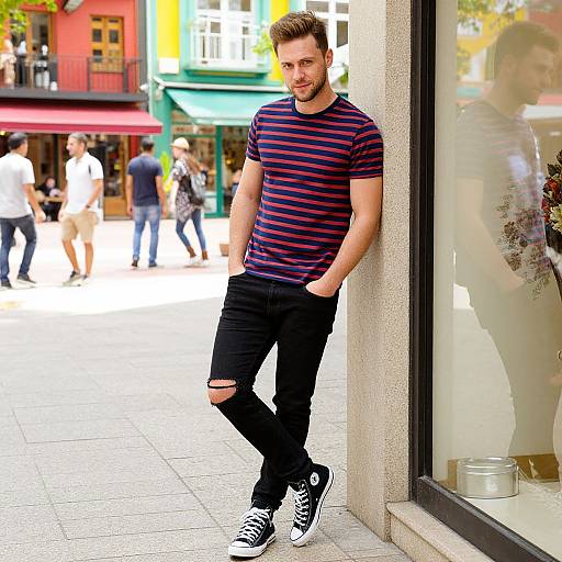 Photograph of a bearded, blond man in a striped shirt and black ripped jeans, leaning against a storefront window in a vibrant, sunlit street
