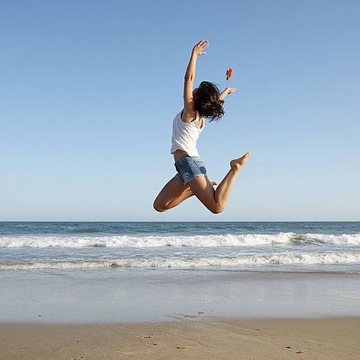 Photograph of a woman with curly black hair, white tank top, and denim shorts, jumping joyfully on a sandy beach with waves crashing in the